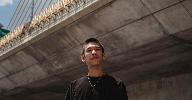 Air Suspension - Young contemplative Asian male athlete with skateboard looking away under cloudy sky in town